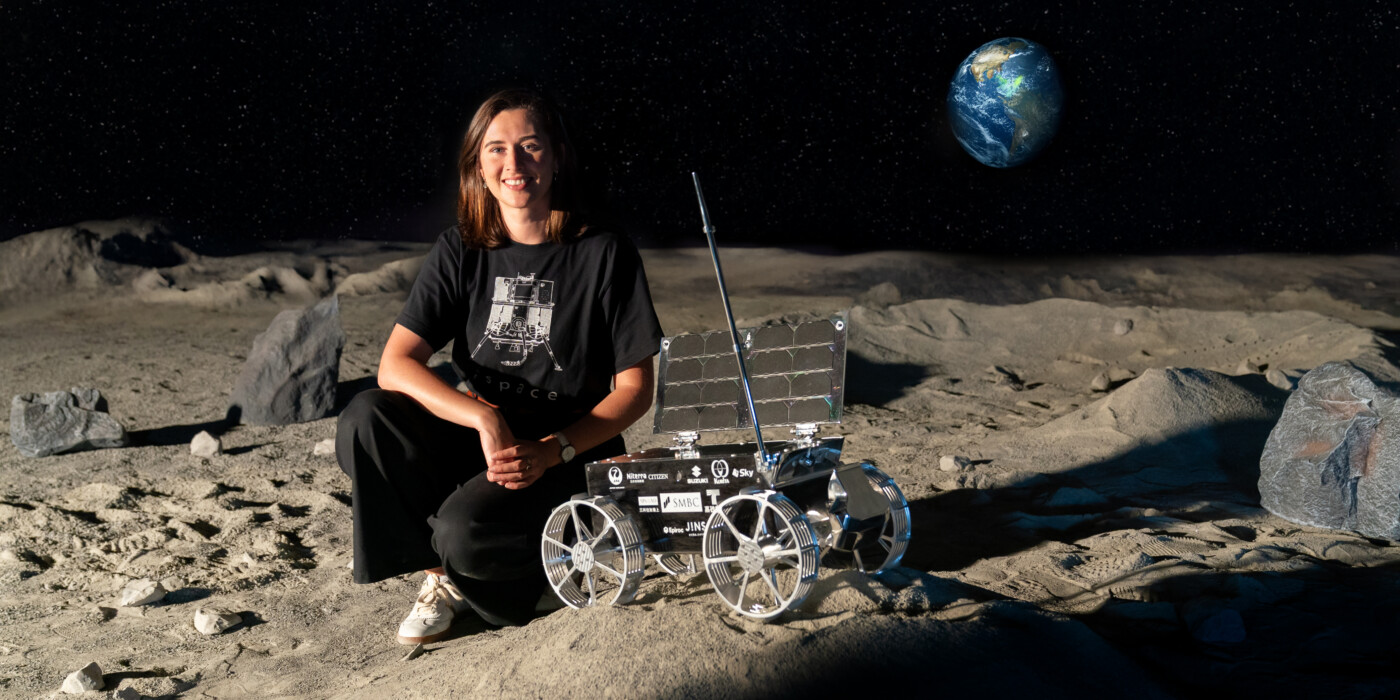 Charlotte Nassey dans l'espace test luniare de la société ispace, accompagnée d'un rover, avec la Terre en arrière plan
