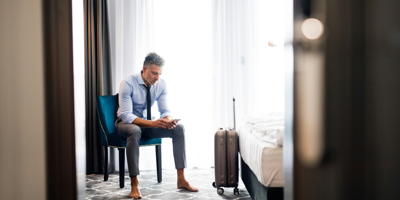 Un homme en costume mais pieds nus, assis dans une chambre d’hôtel avec une valise à roulettes