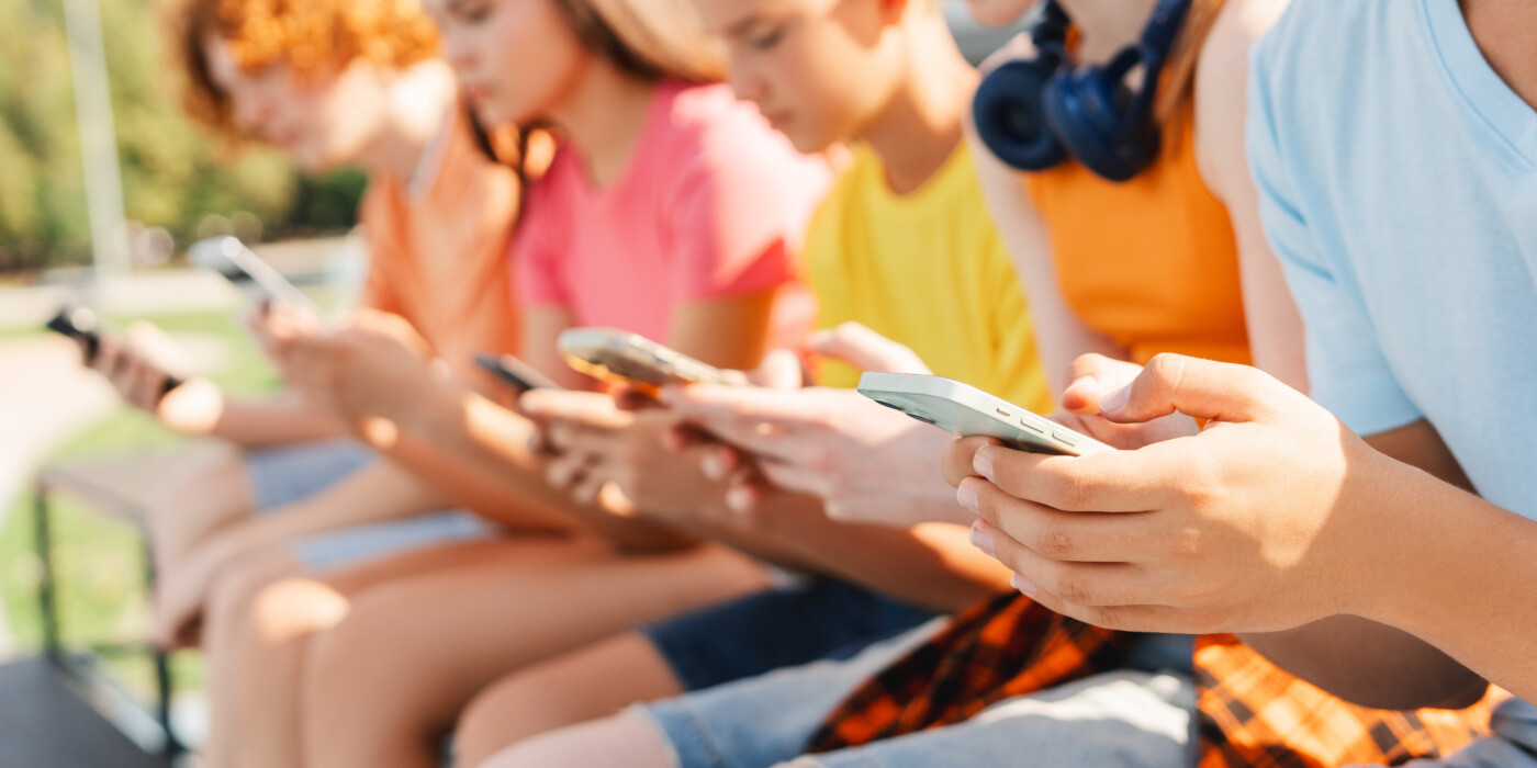 Close up of teenagers holding smartphones sitting in a row on bench on the street. Technology addiction concept