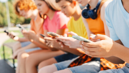 (crédit: Envato) Close up of teenagers holding smartphones sitting in a row on bench on the street. Technology addiction concept