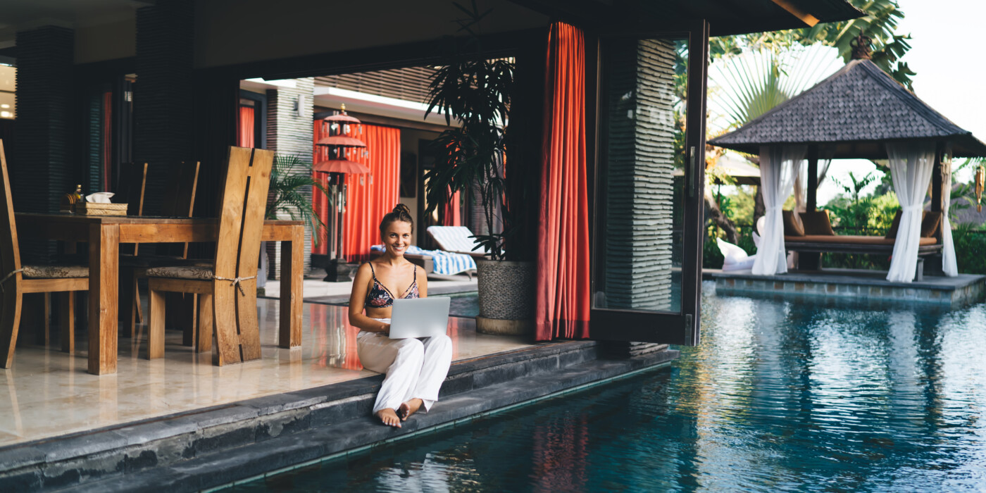 Full body of smiling young female freelancer in white trousers and bra sitting at poolside and working remotely via laptop during vacation