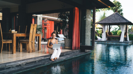 Full body of smiling young female freelancer in white trousers and bra sitting at poolside and working remotely via laptop during vacation
