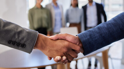 Le mot d'ordre : une transmission ne s'improvise pas. Cette étape requiert des années de préparation (crédit : Envato). Une poignée de mains avec en arrière-plan des salariés dans un bureau